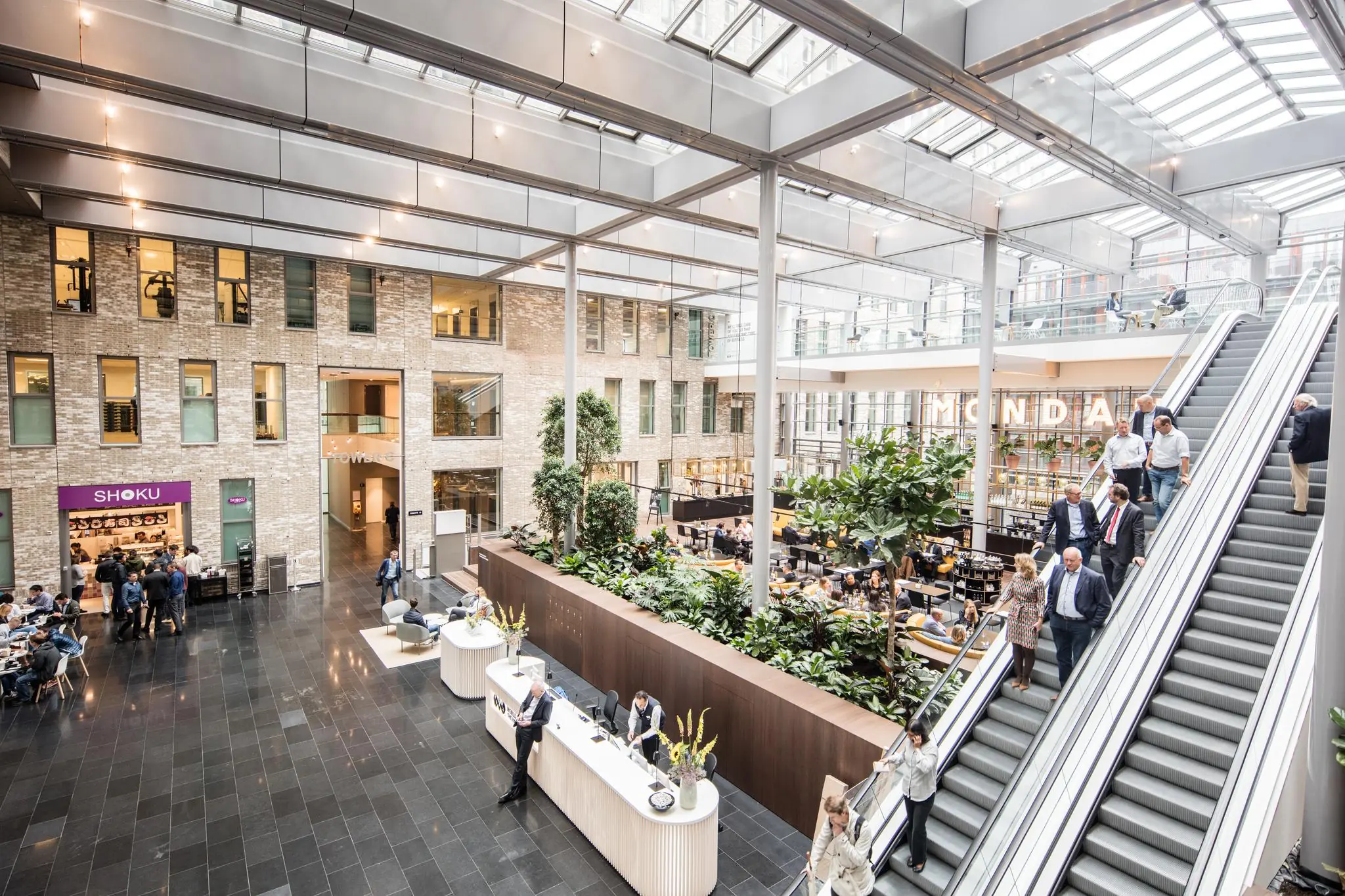 Een moderne, lichte en drukbezochte ontvangsthal aan de Prinses Beatrixlaan met een roltrap, receptiebalie, restaurant en kantoorruimtes.